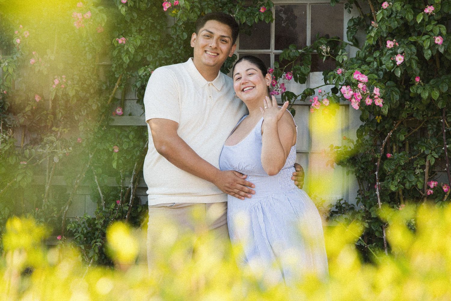A smiling couple embraces in a sunlit garden surrounded by pink roses and yellow flowers.