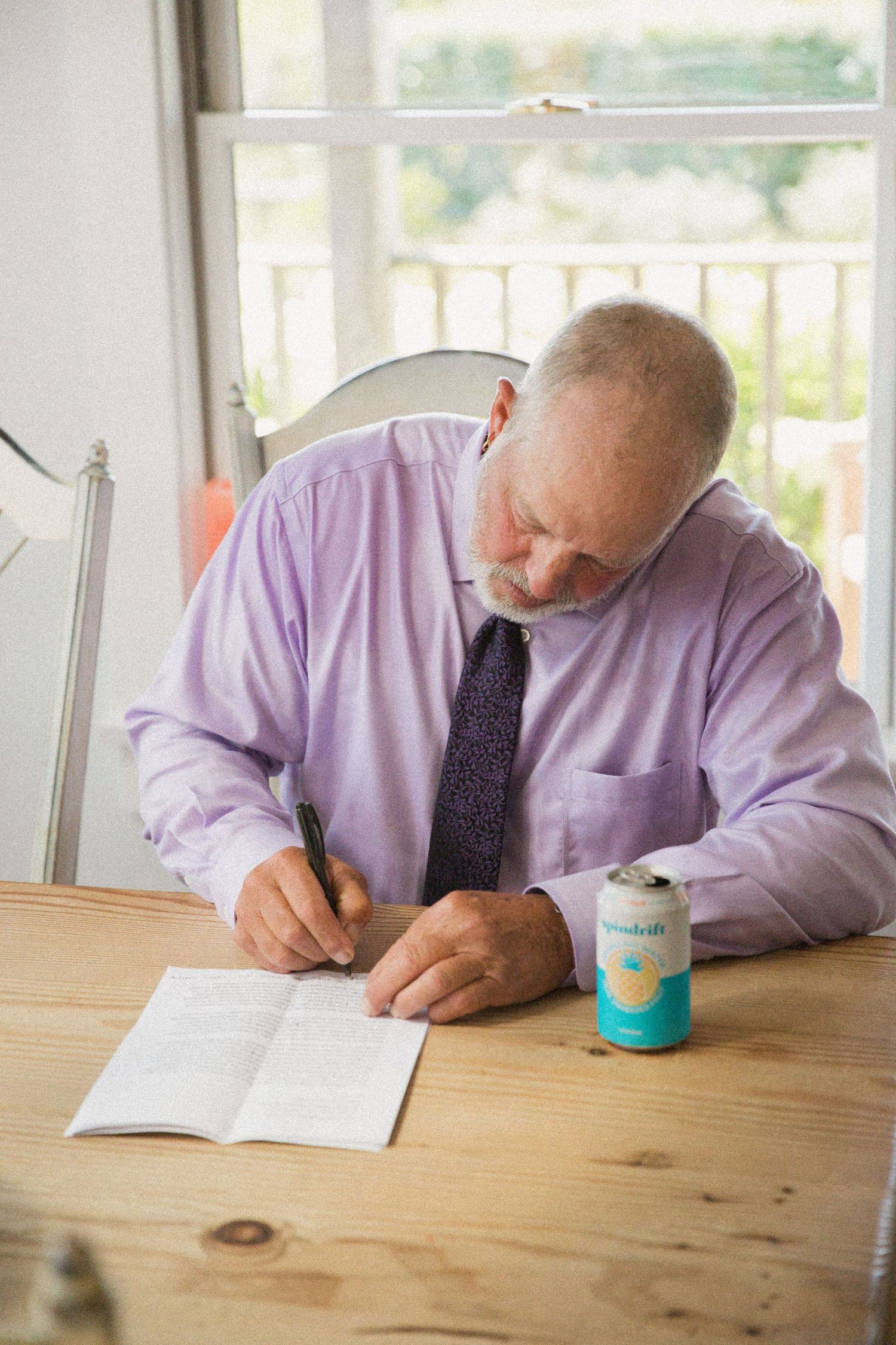 Person in lavender shirt signing documents at wooden table near window.