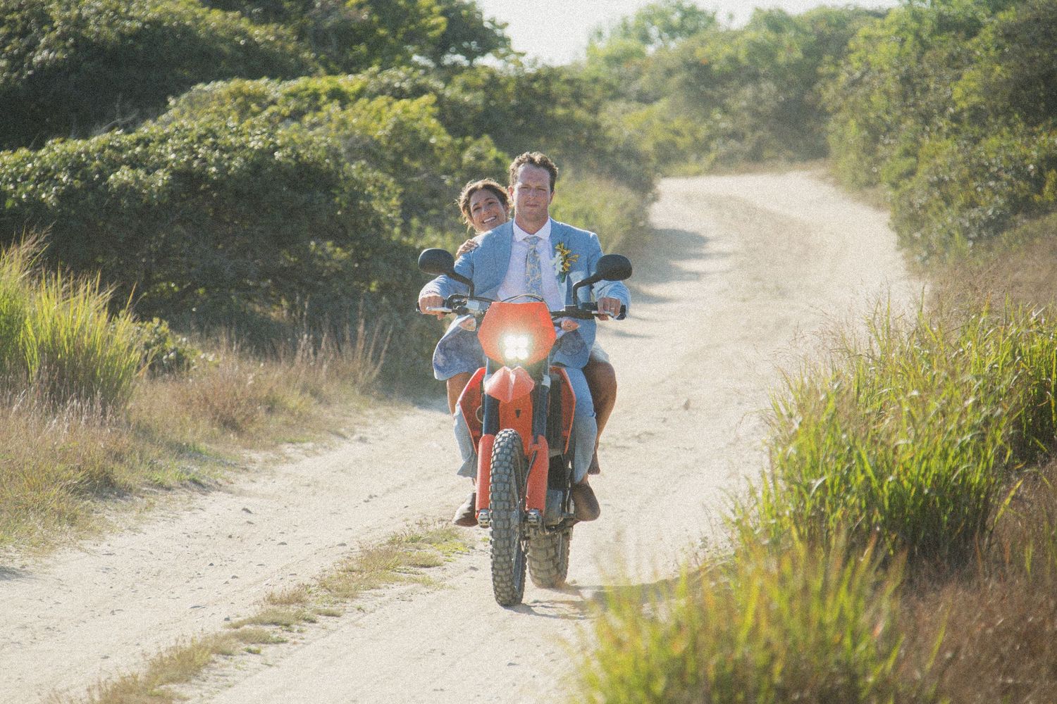 Wedding couple riding orange dirt bike on sandy path through coastal vegetation.