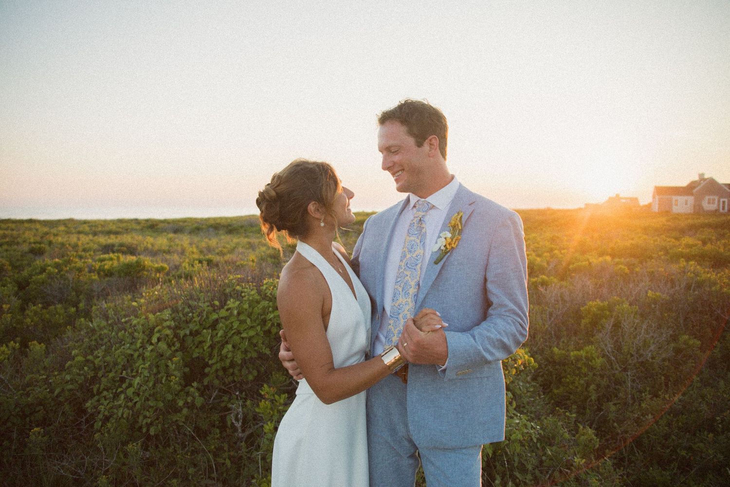 A couple embraces during golden hour with wildflowers and coastal scenery in the background.