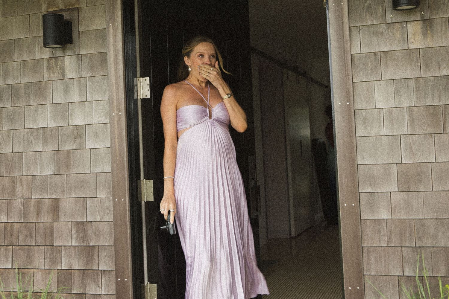 Pregnant woman in lavender pleated strapless gown standing in doorway, covering mouth with hand.