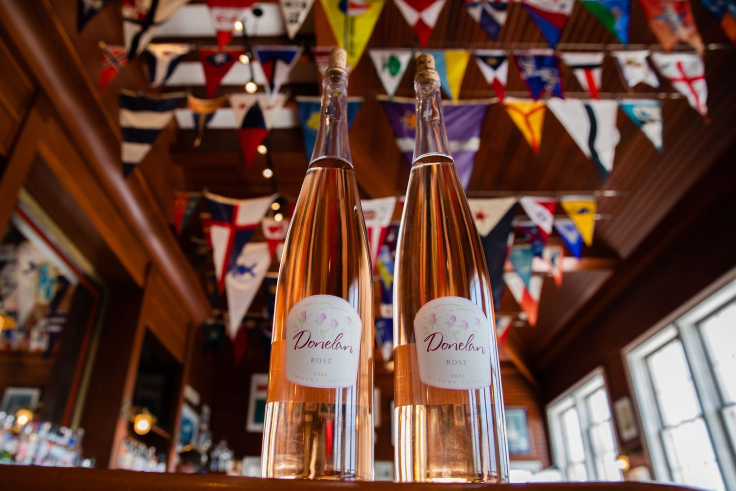 Two elegant bottles of Dorchin rosé wine displayed on a wooden bar with colorful bunting flags in the background.