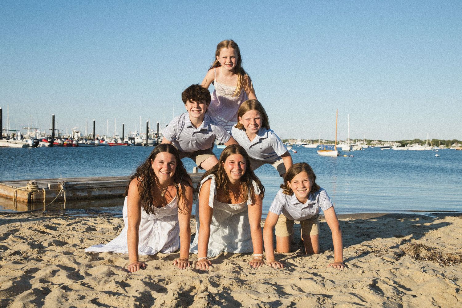 Five children form a human pyramid on a sandy beach with boats and blue water in the background.