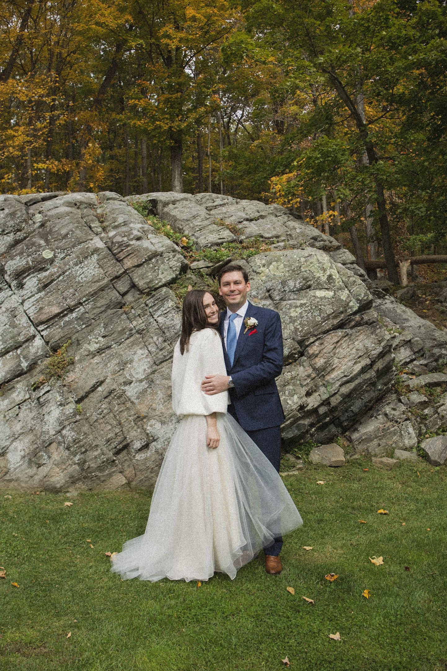 A smiling bride in a tulle skirt and white shawl embraces her groom in a navy suit before a large rock and autumn trees.