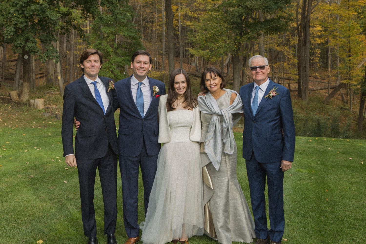 Five people in formal attire, including a bride in a white gown, pose together on a grassy lawn surrounded by autumn trees.