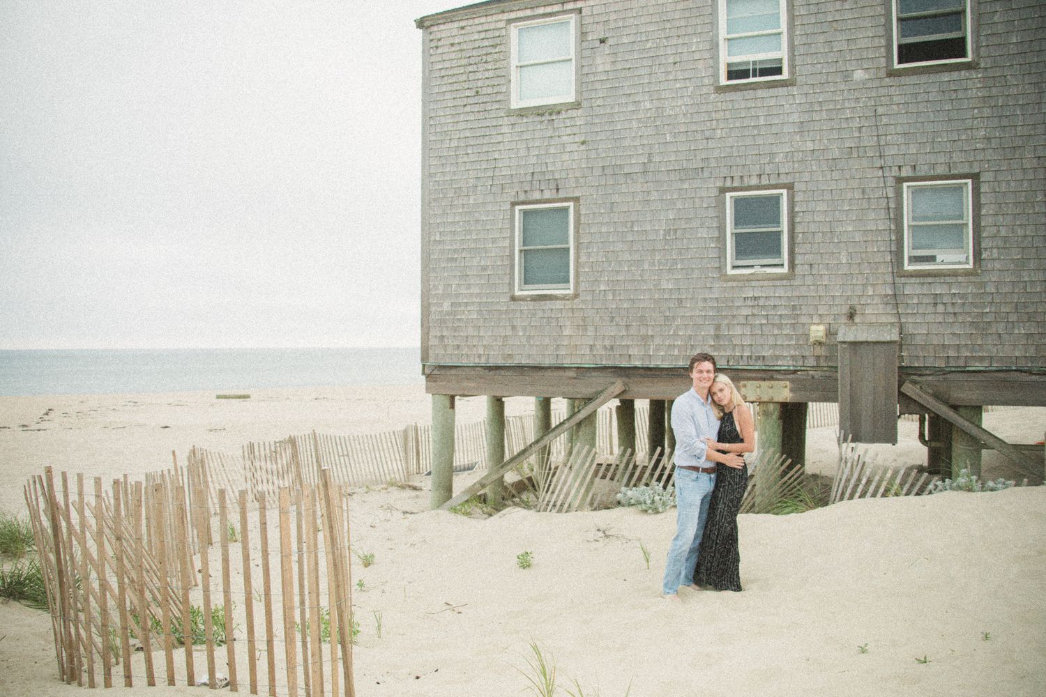 A weathered gray shingled house sits among beach dunes with wooden fencing.