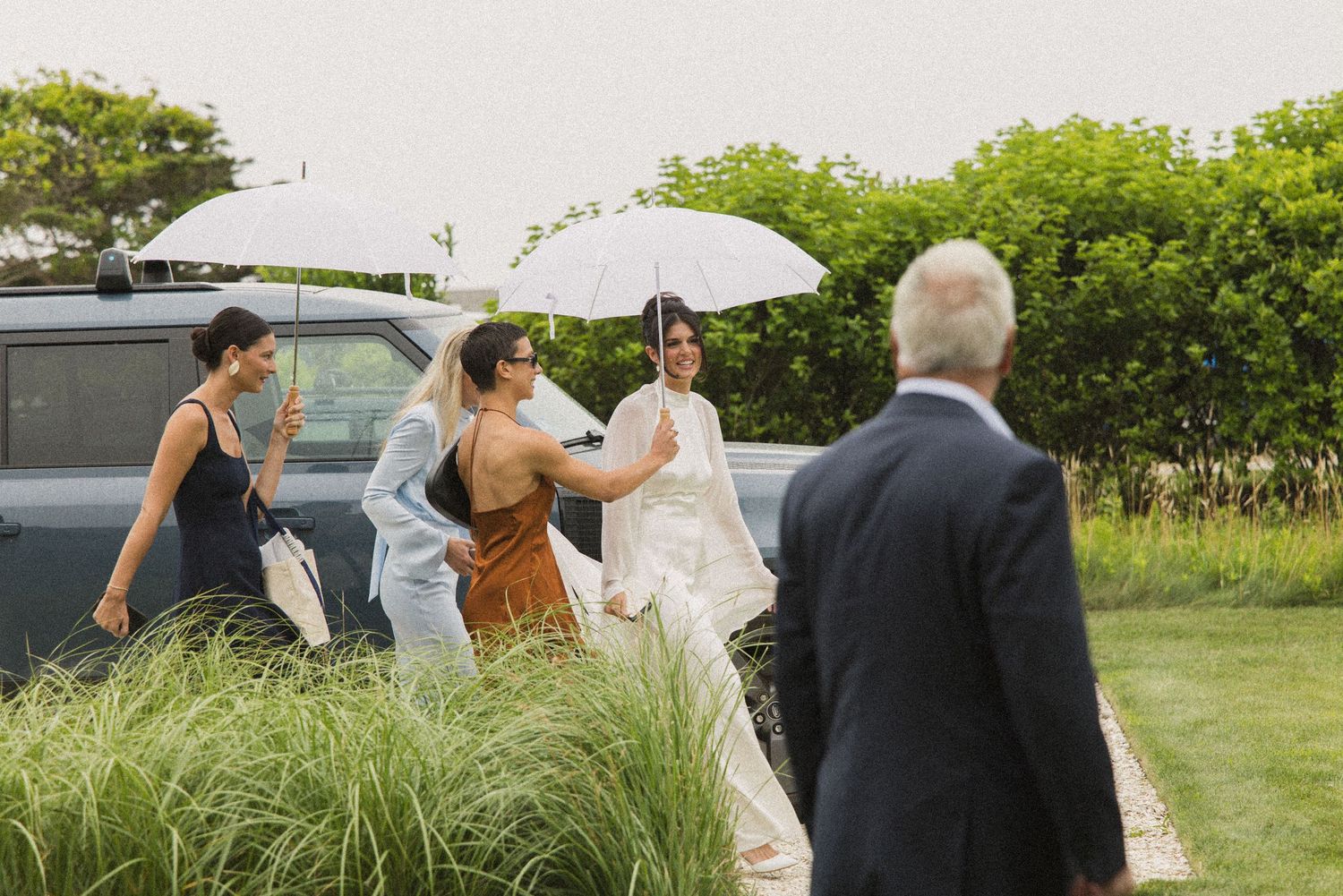 Bride in white gown with umbrella walks with wedding party toward vintage car on rainy outdoor venue.