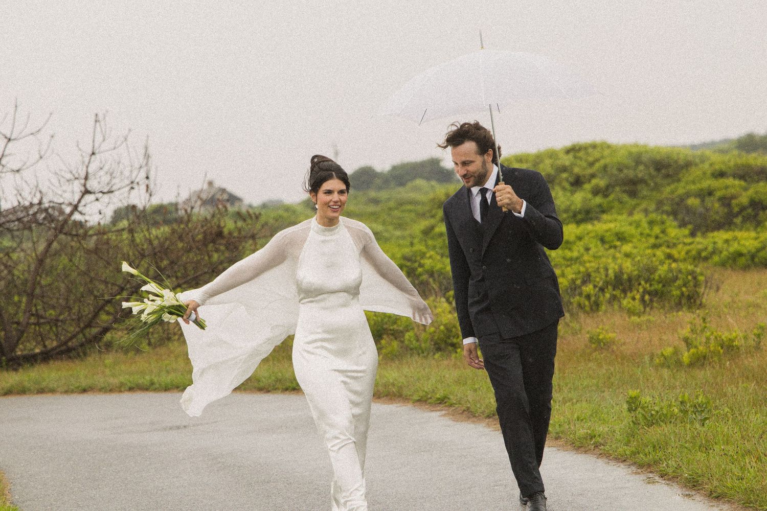 A bride and groom walk on a rainy day path, she holds flowers while he carries a white umbrella outdoors.
