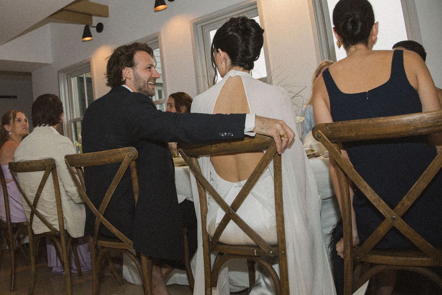 Elegant wedding reception with guests in formal attire seated at wooden cross-back chairs in a bright venue.