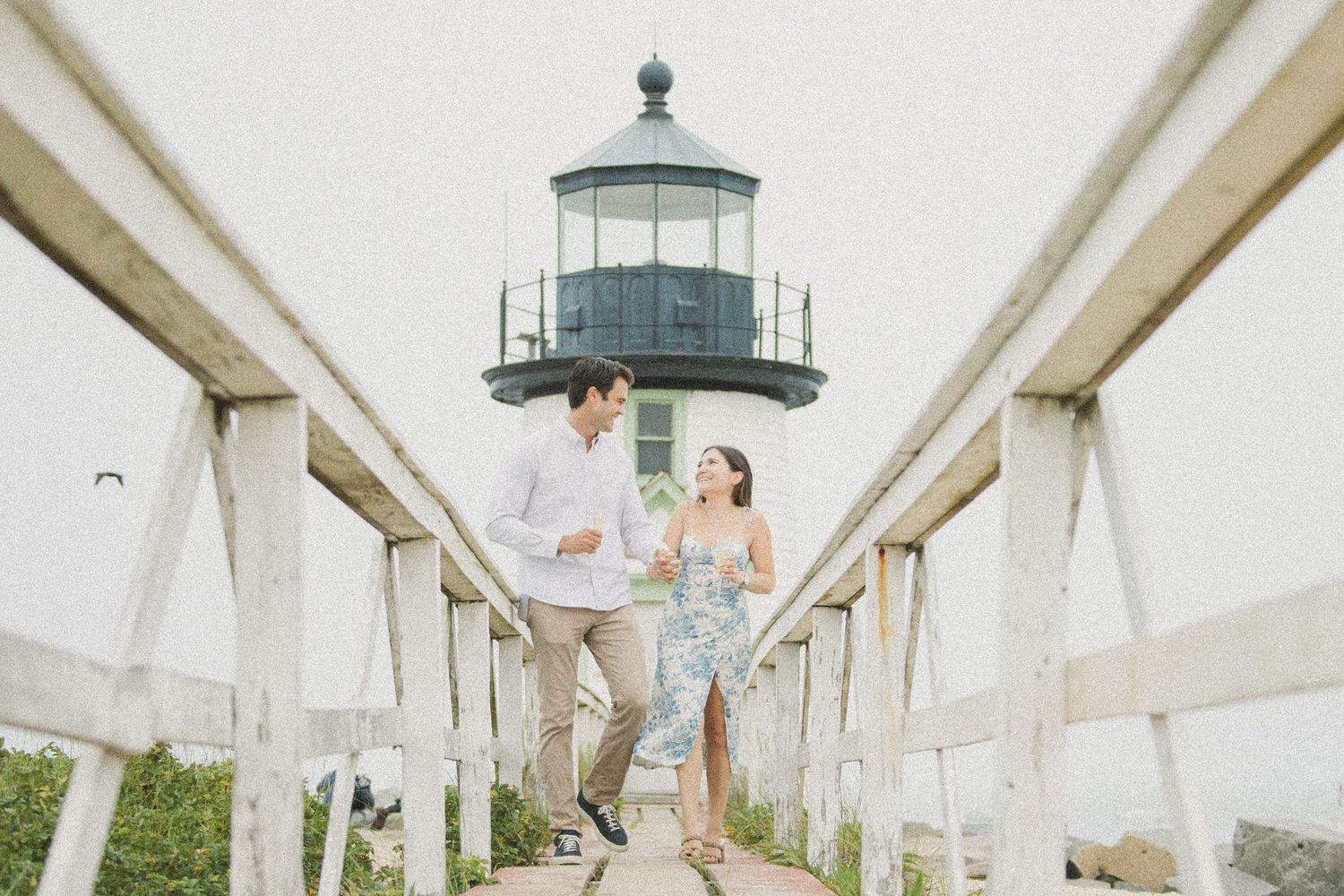 Couple holding hands on a wooden bridge walkway with a classic lighthouse backdrop on a misty day.
