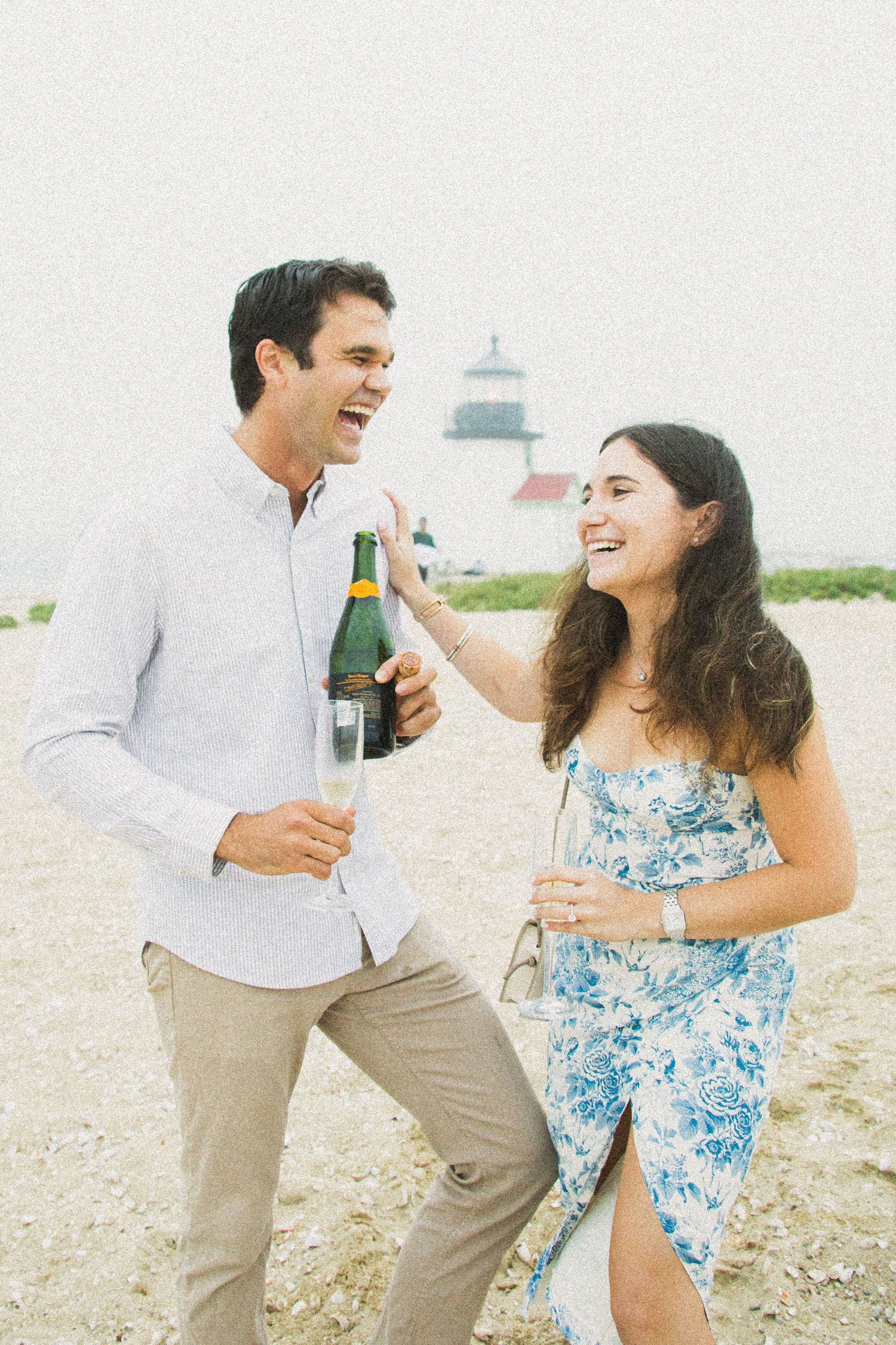 Happy couple celebrating on a beach with champagne, lighthouse in background, woman in floral dress, man in white shirt.