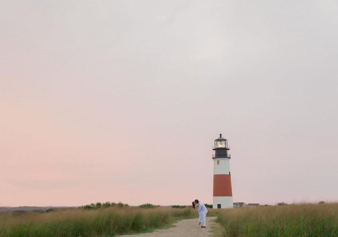 A couple embraces on a path leading to a red and white lighthouse at sunset on Nantucket.