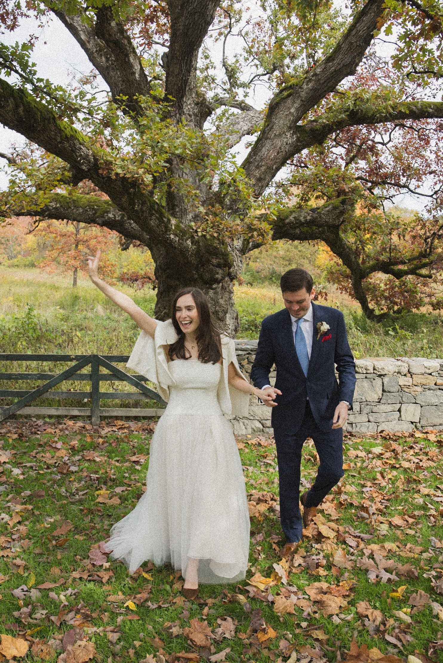 A joyful bride in a white tulle gown raises her arm while holding hands with her groom on an autumn lawn beneath a large old oak tree.