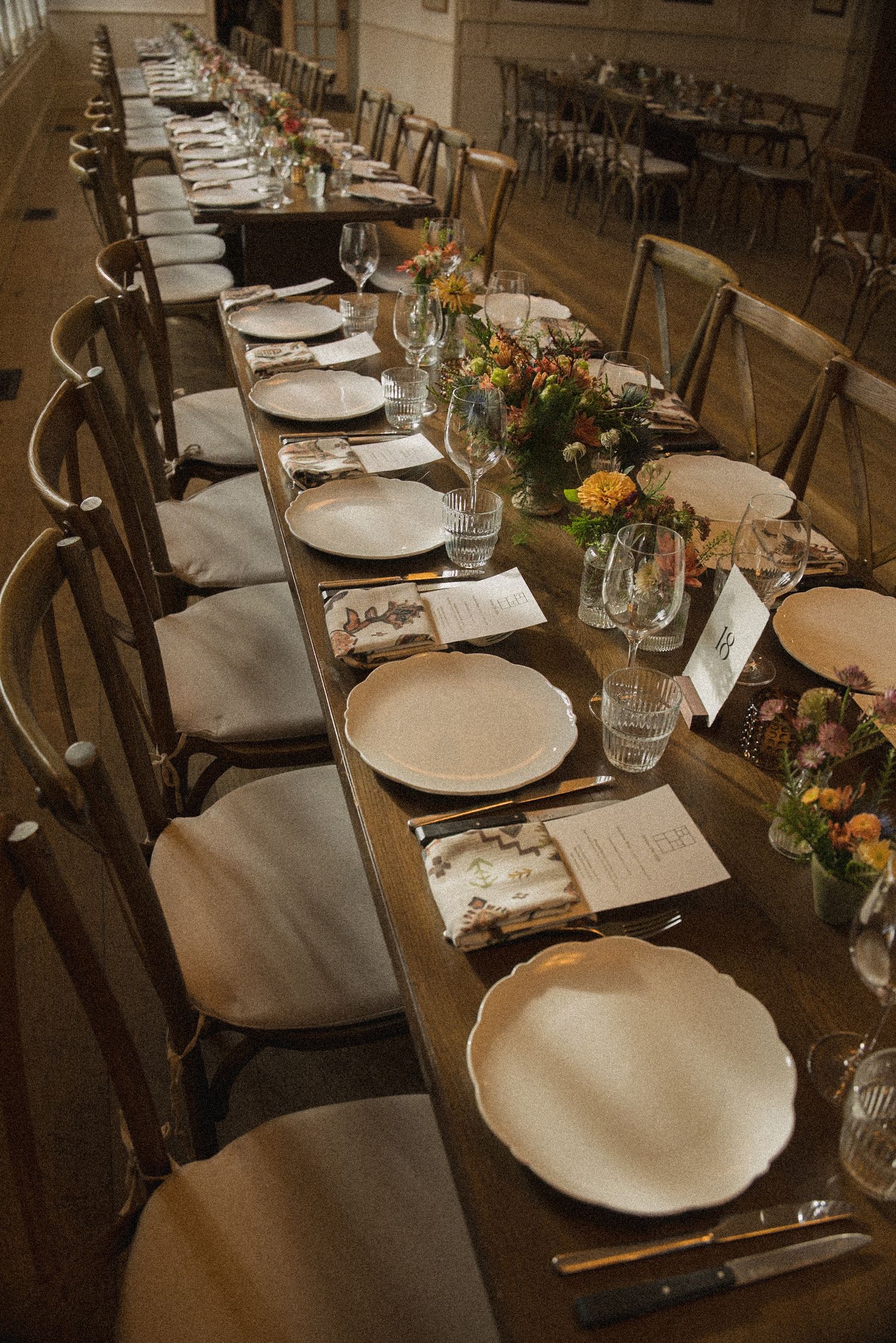 Long rustic wooden banquet table set with white plates, folded menus, glassware and colorful floral centerpieces beside cross-back chairs.