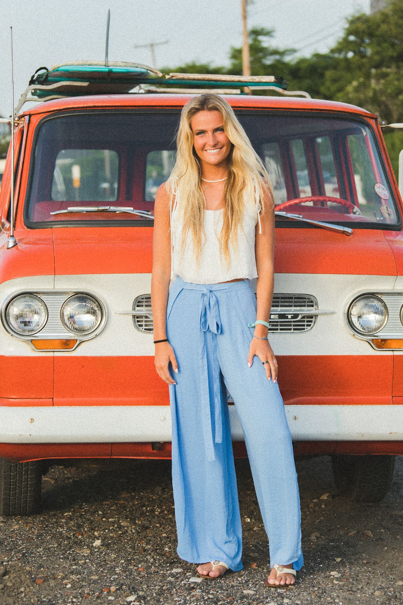 Smiling blonde woman in blue wide-leg pants and white top posing in front of a vintage red van with surfboard on top.