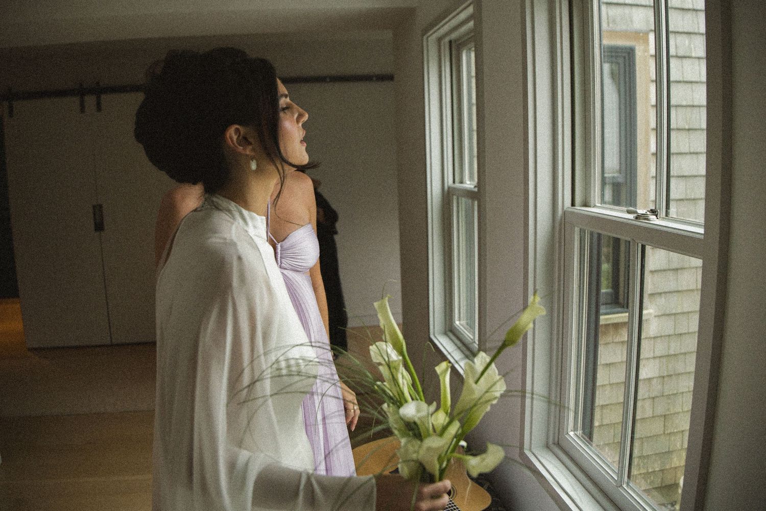 Two women in lingerie stand by a window, one holding white tulips, in a softly lit room.