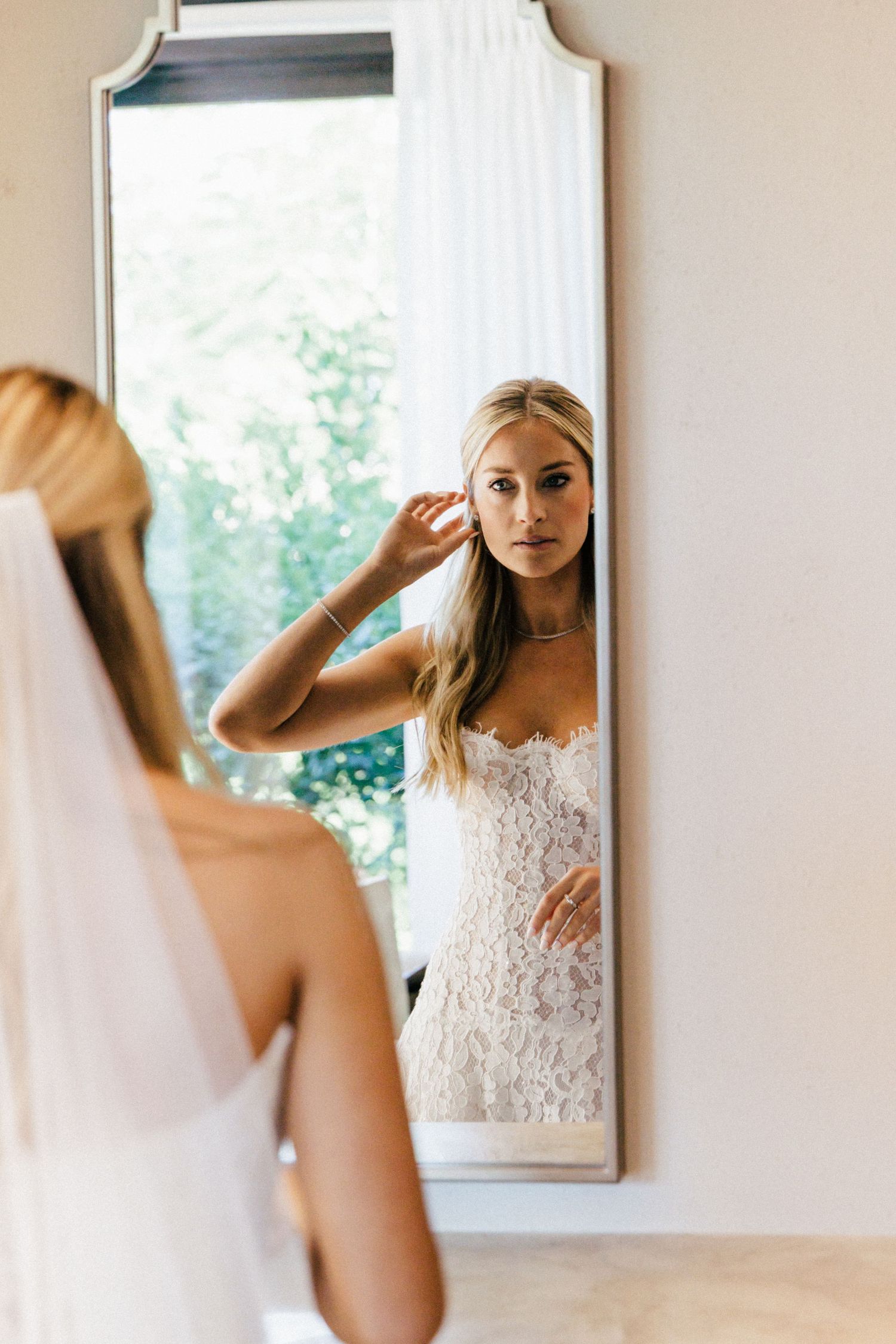 Bride looking in mirrors while wearing white lace wedding gown in bright interior space.