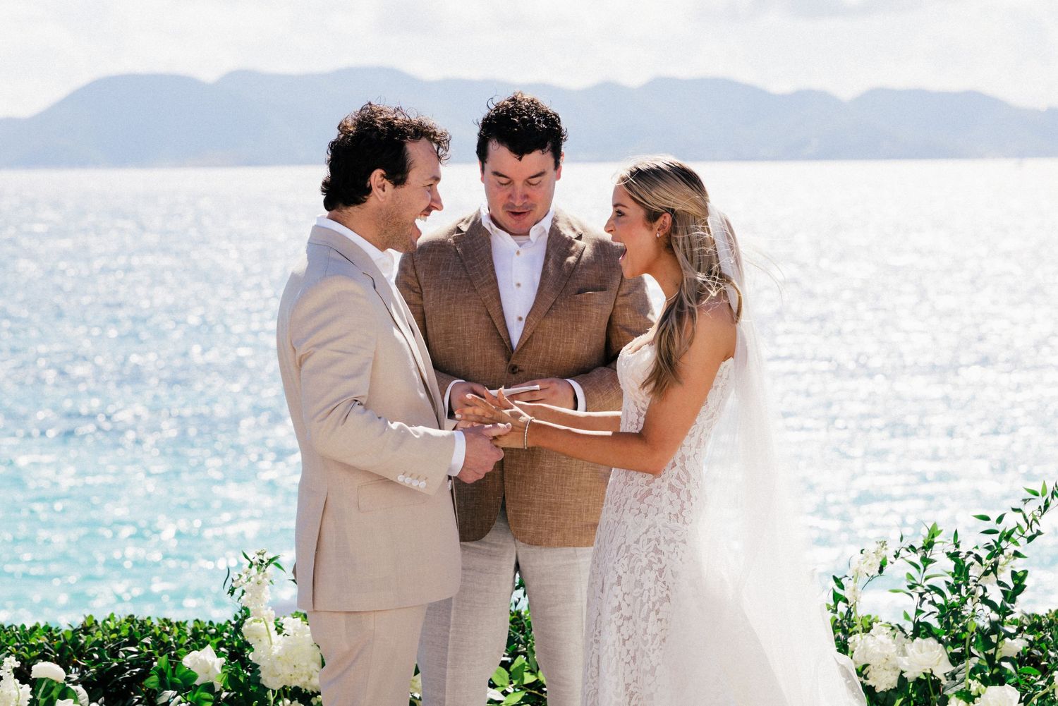 Coastal wedding ceremony with elegant white floral arrangements and mountains in the distance.