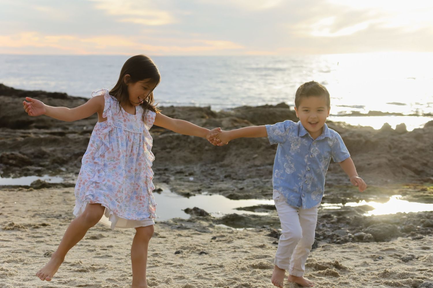 Two children hold hands while playing and running on a rocky beach at sunset.