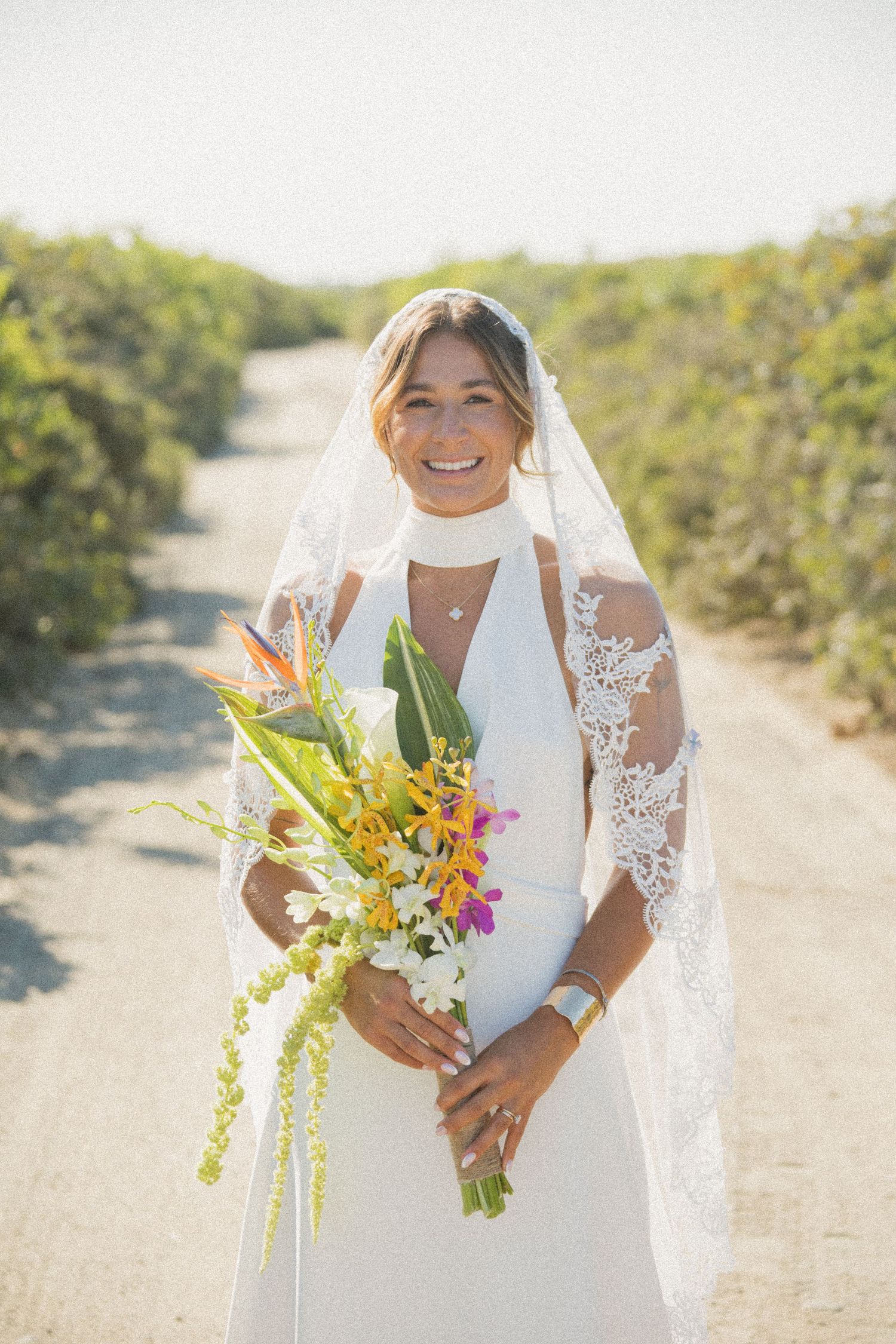 Bride in lace detailed dress holds a colorful tropical bouquet on a sunny outdoor path.