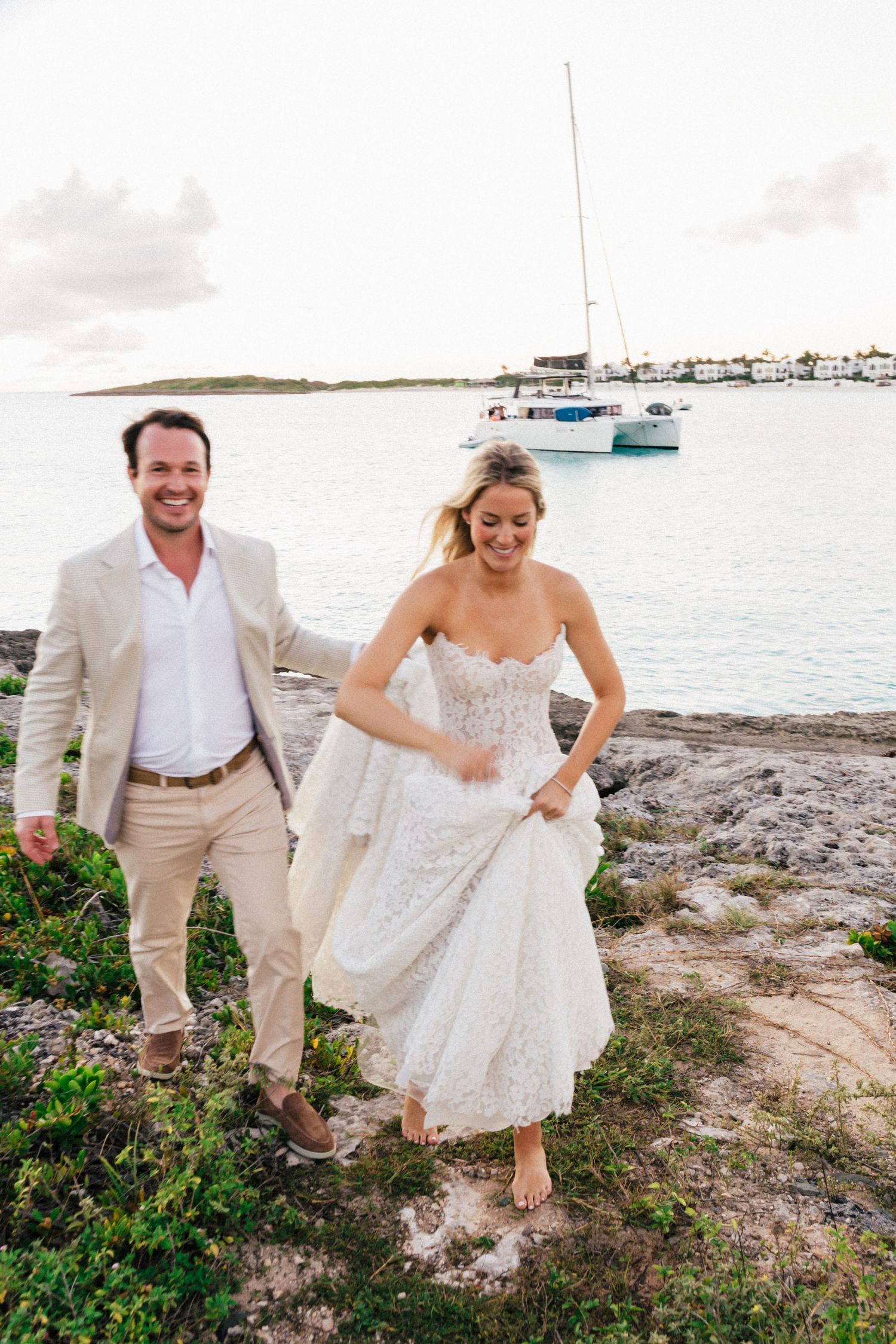 Wedding couple walks along rocky coastal path with boats and harbor in background.