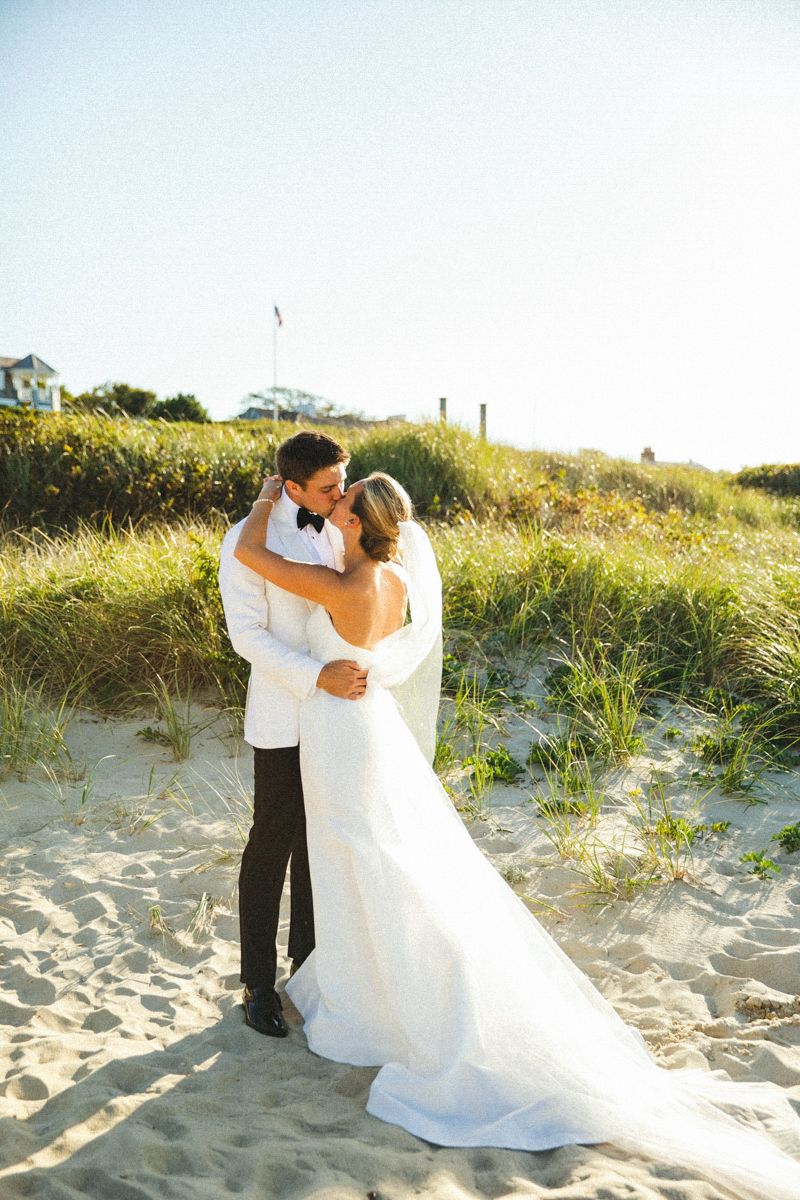 A bride and groom share an intimate moment on a sandy beach with coastal dunes in the background.