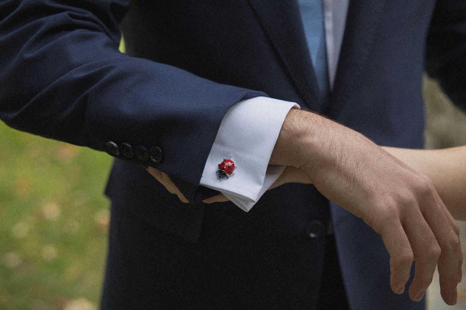 Close-up of a groom's wrist showing a white dress shirt cuff fastened with a red ladybug cufflink under a navy suit sleeve.