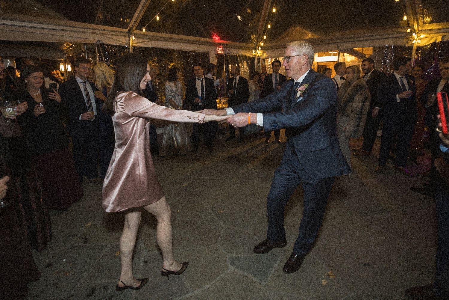 A woman in a blush satin dress and a man in a navy suit hold hands and dance while wedding guests watch under a tent.