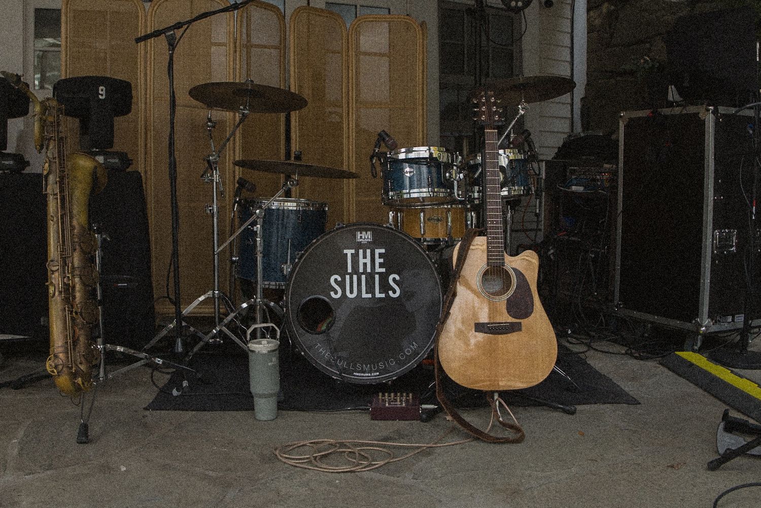 Drum kit with a bass drum labeled The Sulls, an acoustic guitar, and a saxophone set up on a rehearsal stage.