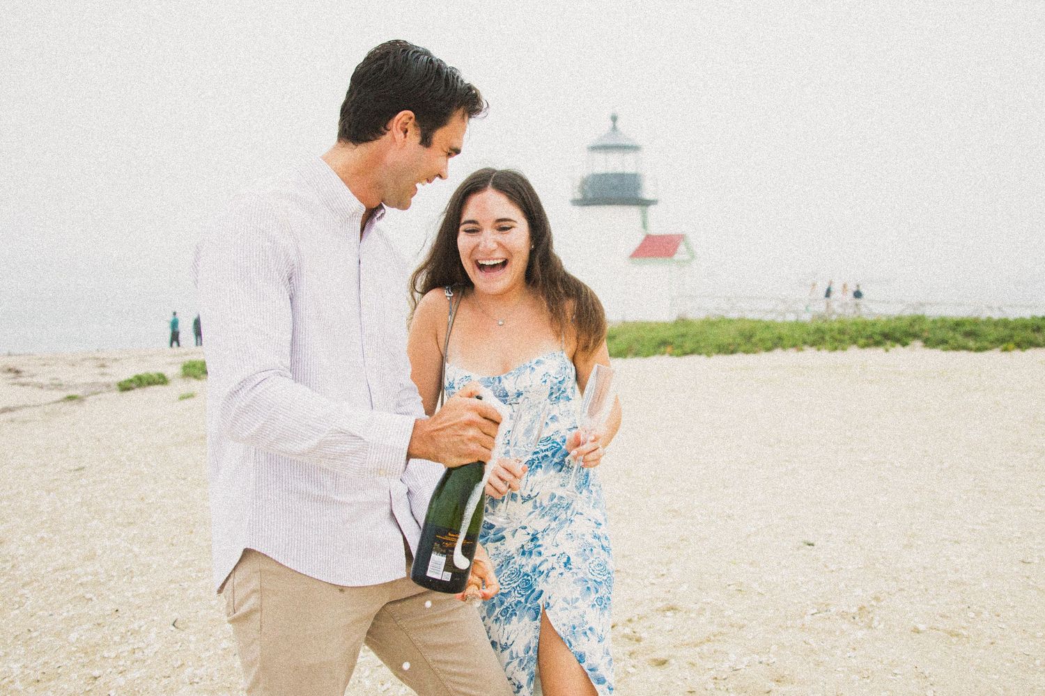 Couple laughing on beach with champagne bottle, lighthouse in background, celebrating engagement.