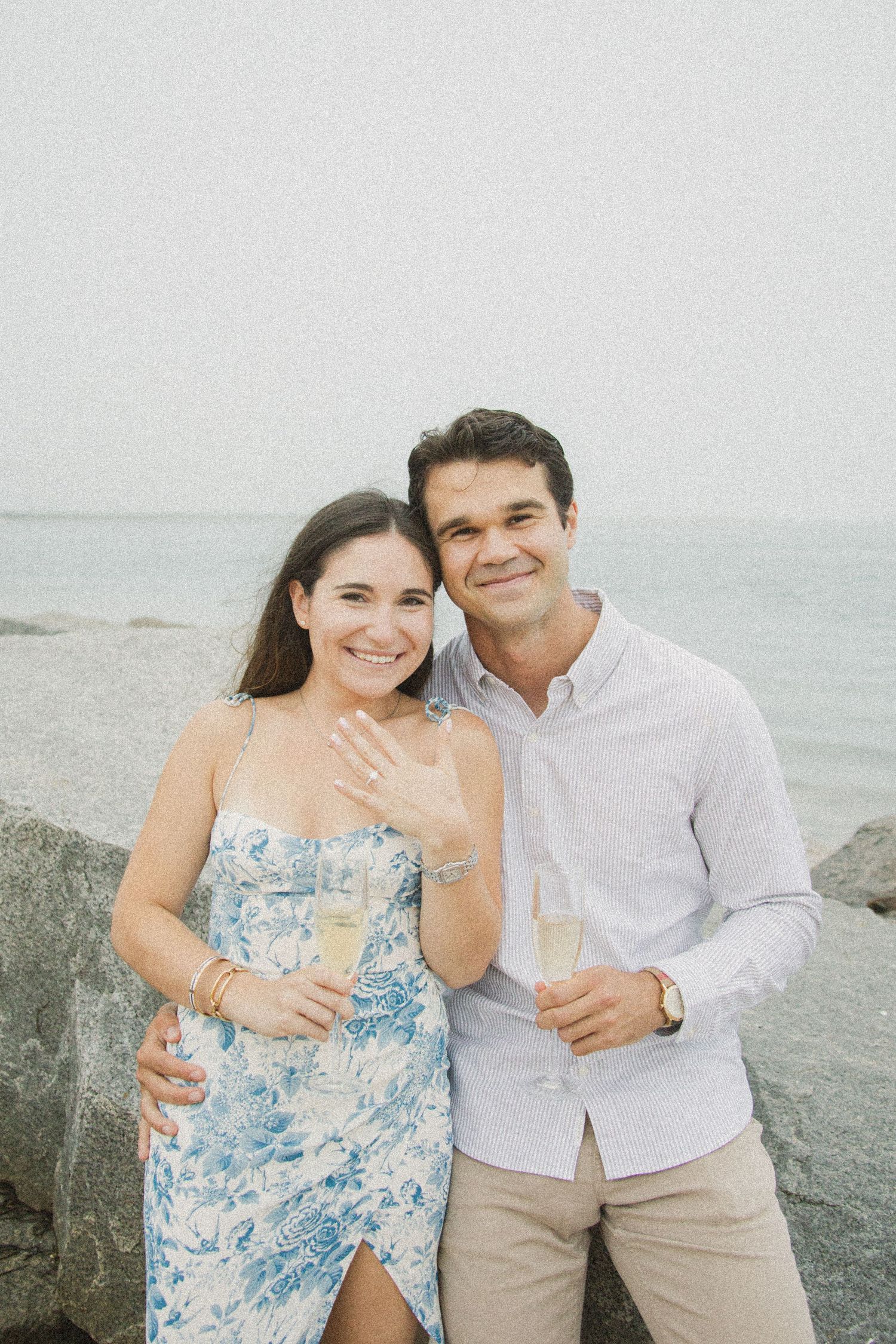 Happy couple celebrating engagement at beach, woman showing ring while holding champagne glasses near rocky shoreline.