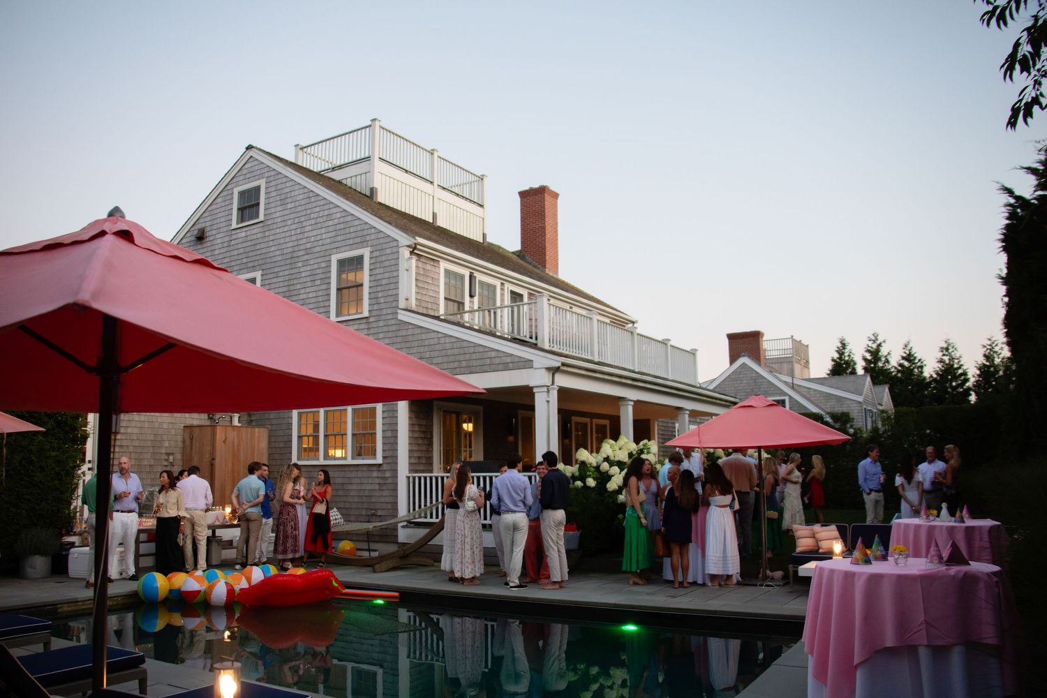 Outdoor evening party by a pool at a gray shingled home, guests mingling under red umbrellas at dusk.