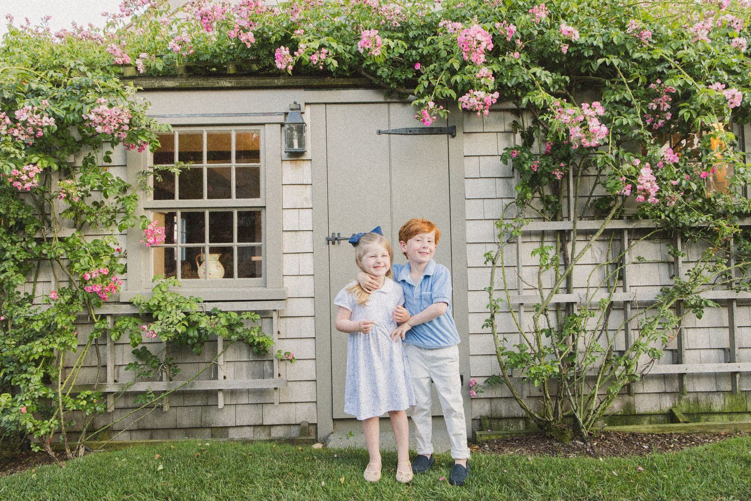 Two children hugging and smiling in front of a charming cottage covered in pink climbing roses.