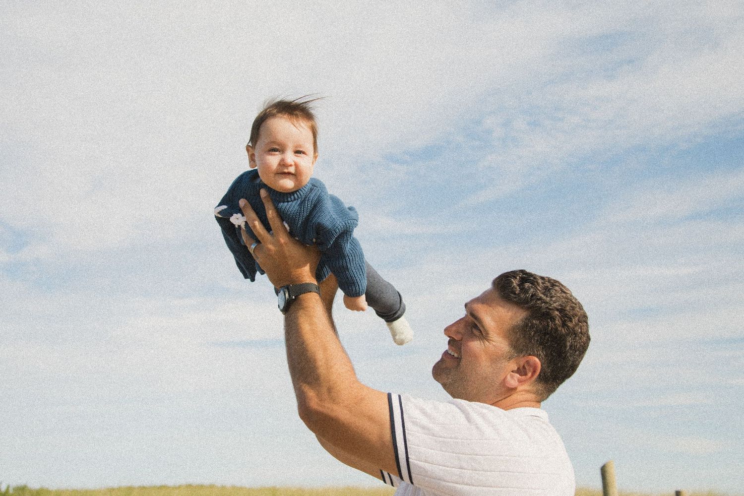 A playful moment captured as an adult lifts a small child high into the air against a blue sky.