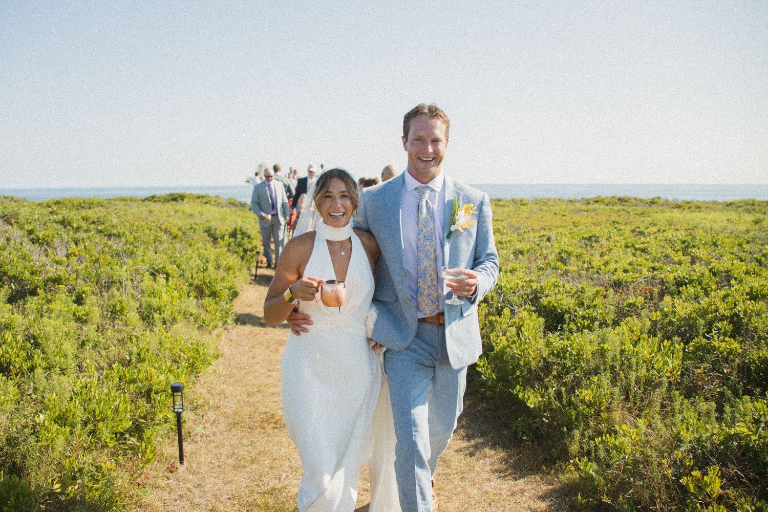 A couple in wedding attire walks along a scenic dirt path surrounded by green foliage on a sunny day.