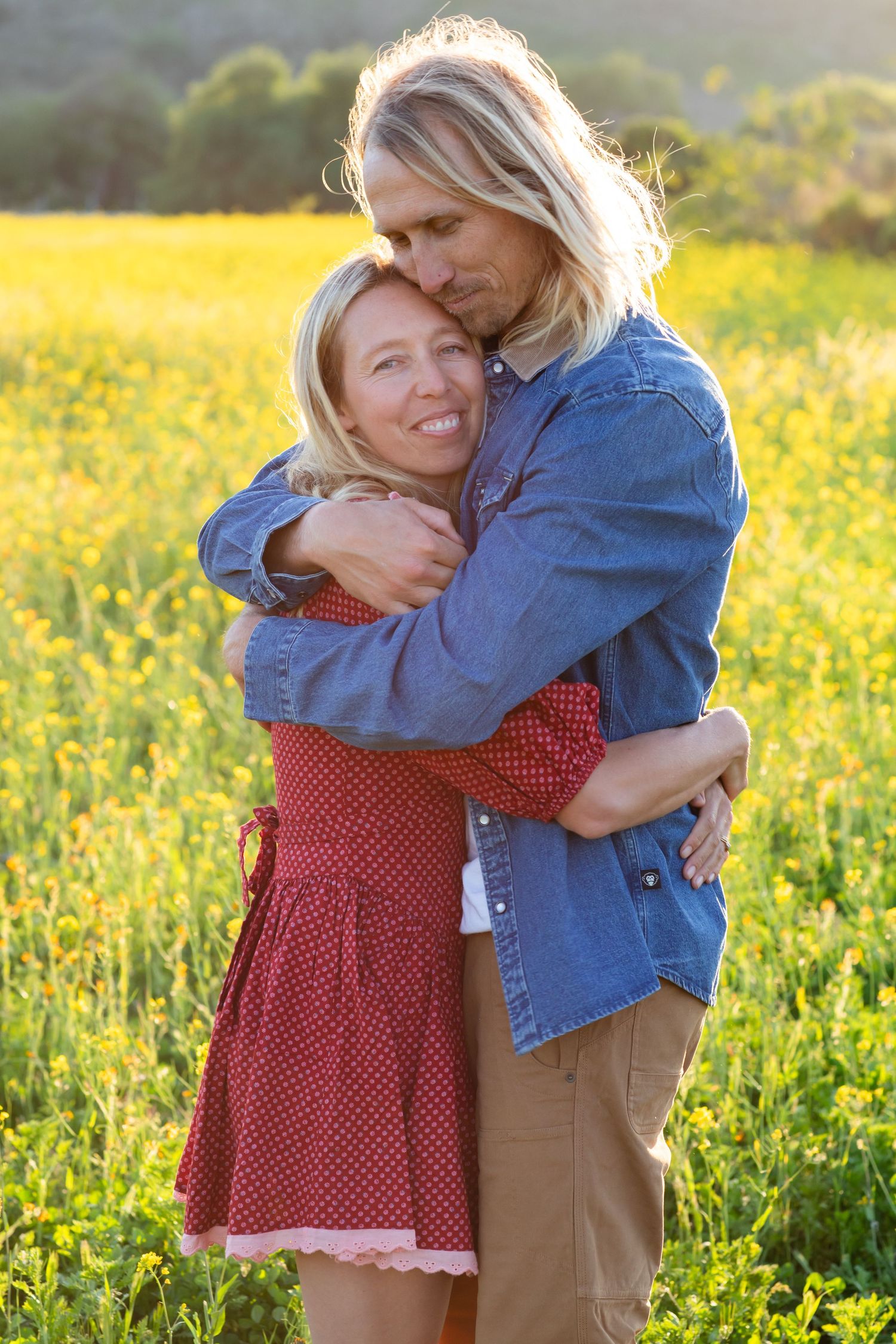 Two people share an embrace while standing in a field of yellow wildflowers at golden hour.
