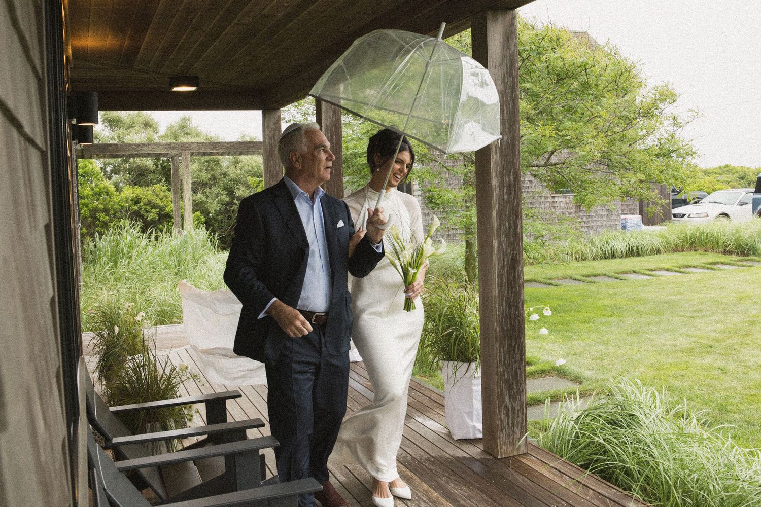 Couple walking on a wooden deck in the rain, woman holding a clear umbrella, surrounded by lush green gardens.