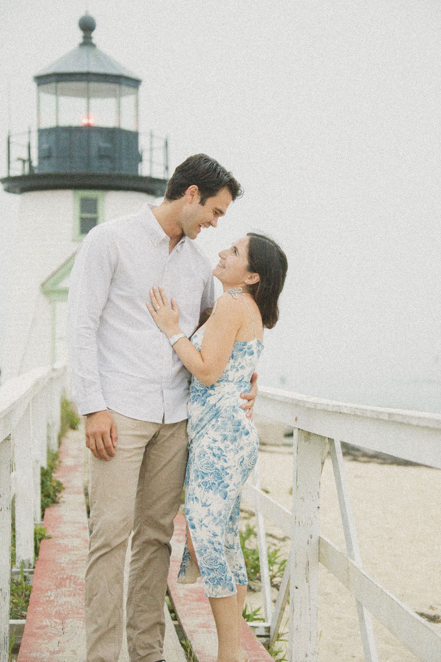 Couple embracing near a lighthouse on a coastal boardwalk, woman in floral dress and man in white shirt, sharing a romantic moment.
