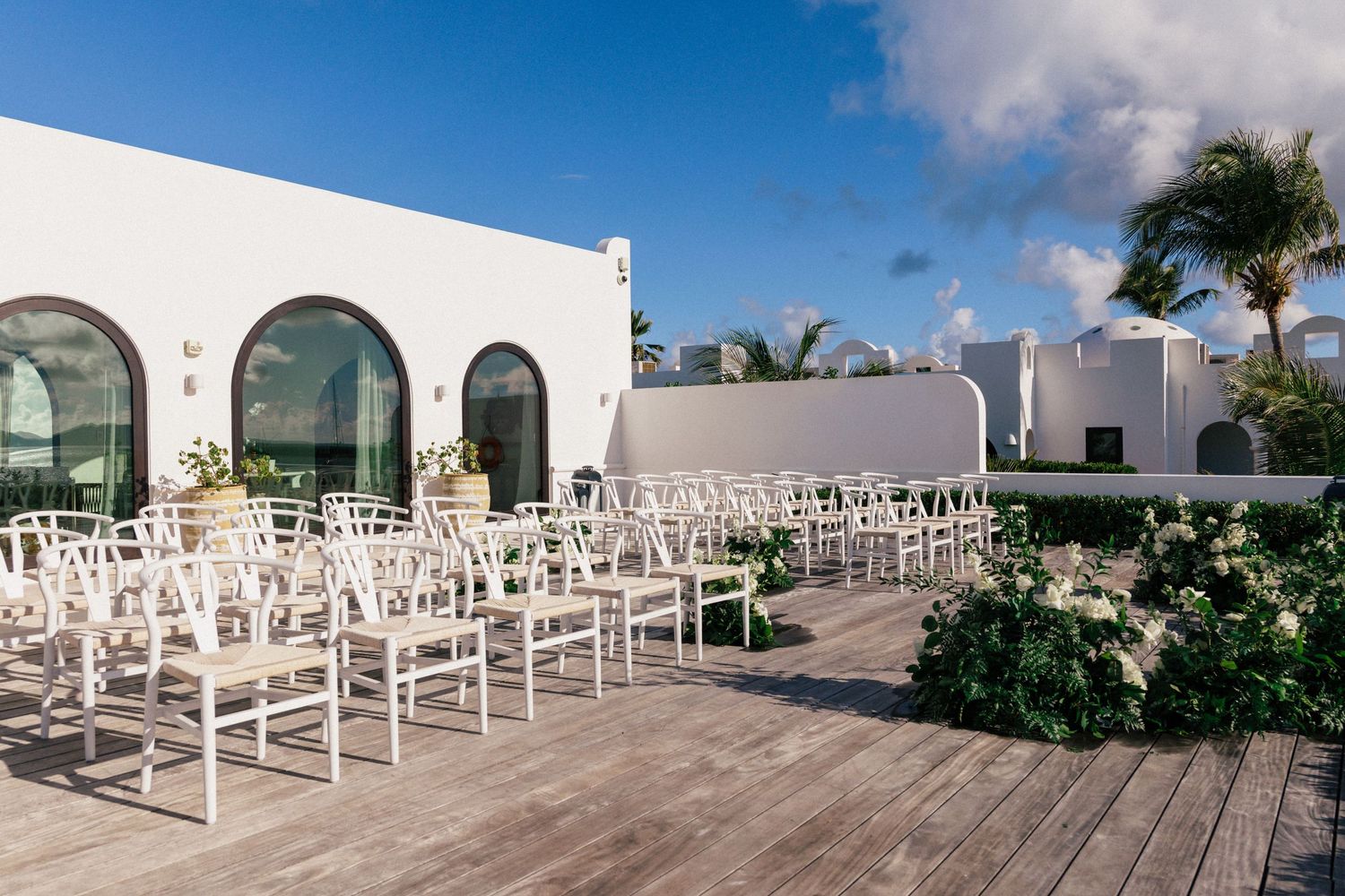 Outdoor wedding ceremony setup with white chairs and arched windows at beachfront venue.