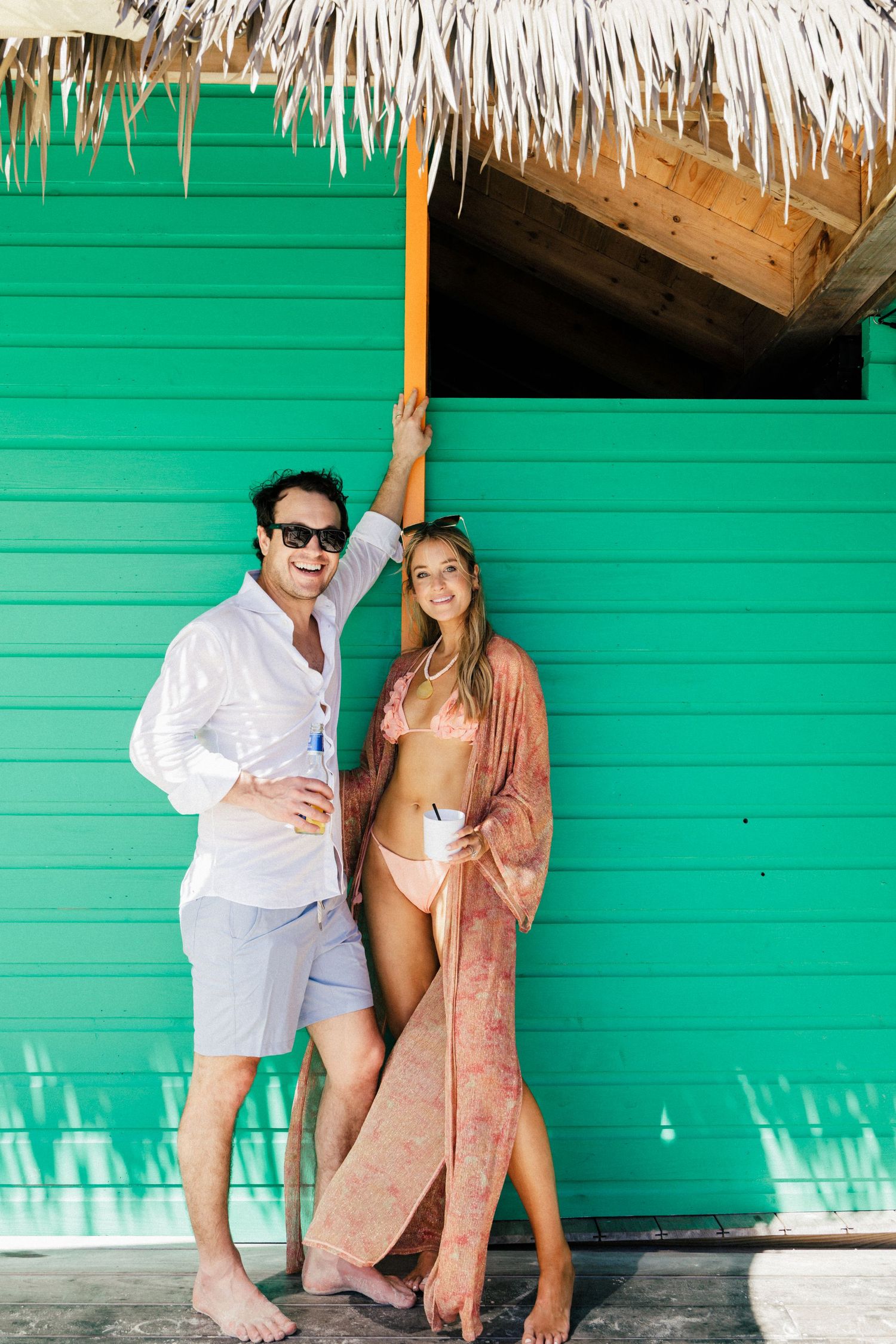 Stylish couple in resort wear standing against vibrant green wall under thatched roof.