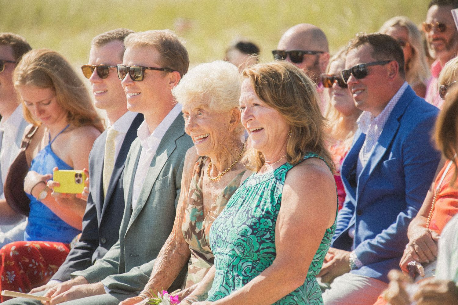 Wedding guests seated in colorful attire watch beachside ceremony.