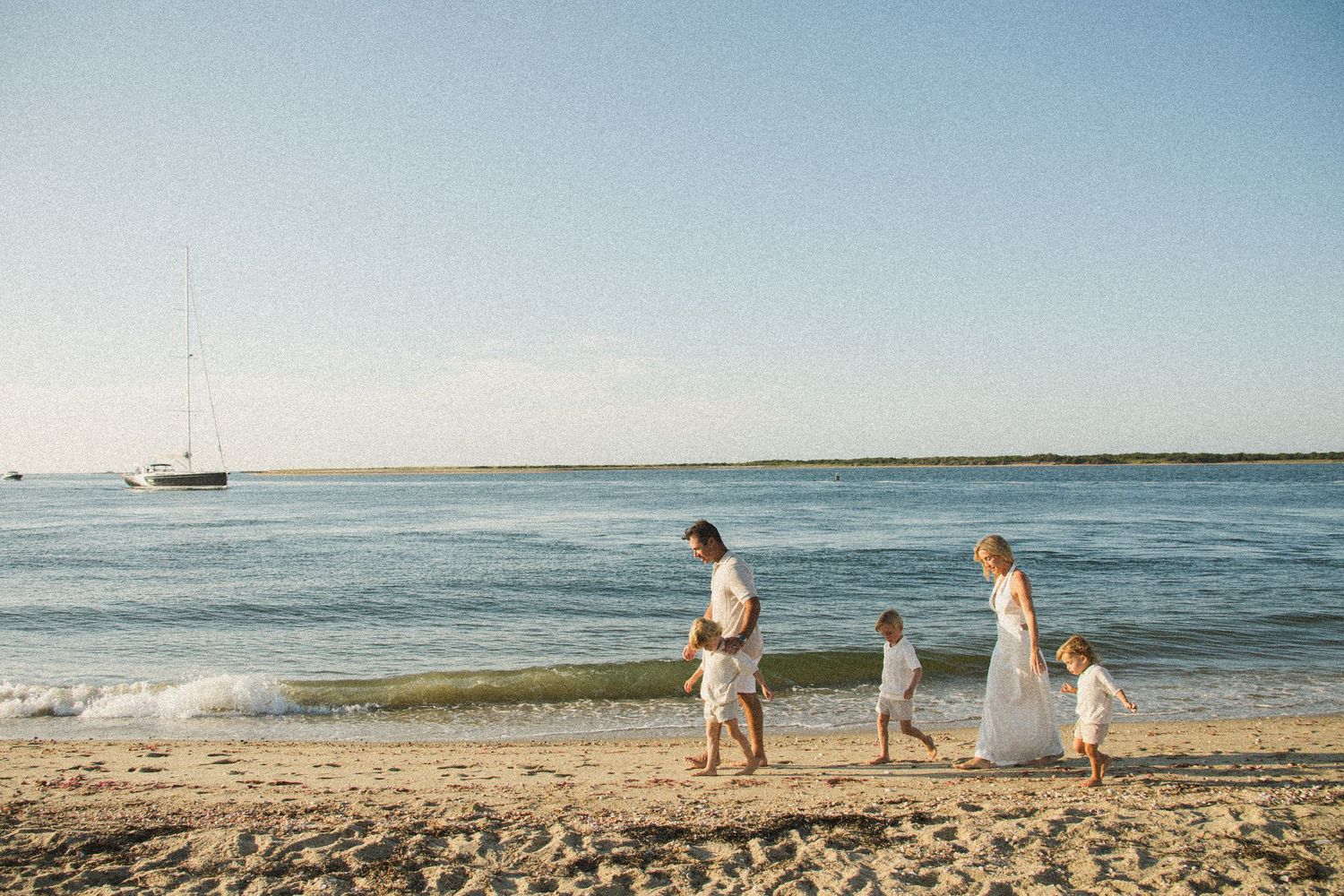 A family walks along a beach shoreline with sailboats visible in the distance.