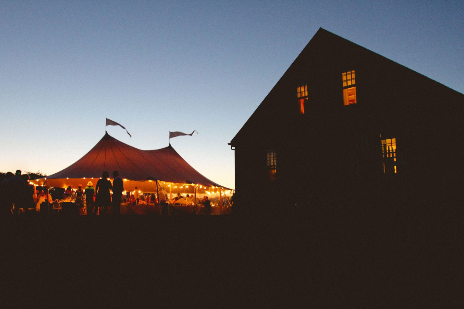 A rustic barn venue glows warmly at dusk with string lights illuminating the interior through windows and doorways.