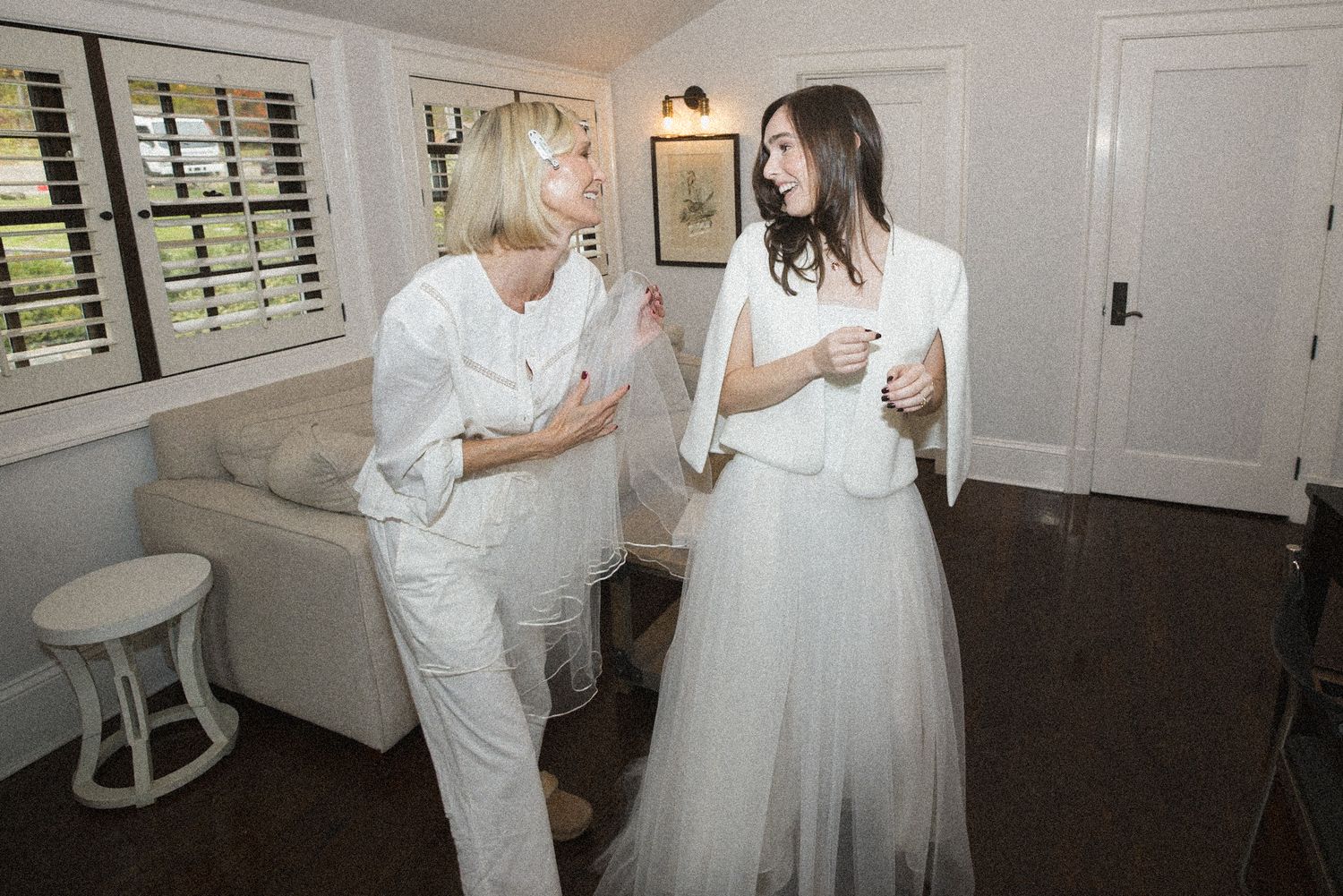 Two women in white outfits stand and talk in a bright room with shuttered windows, a cream sofa and a white side table.