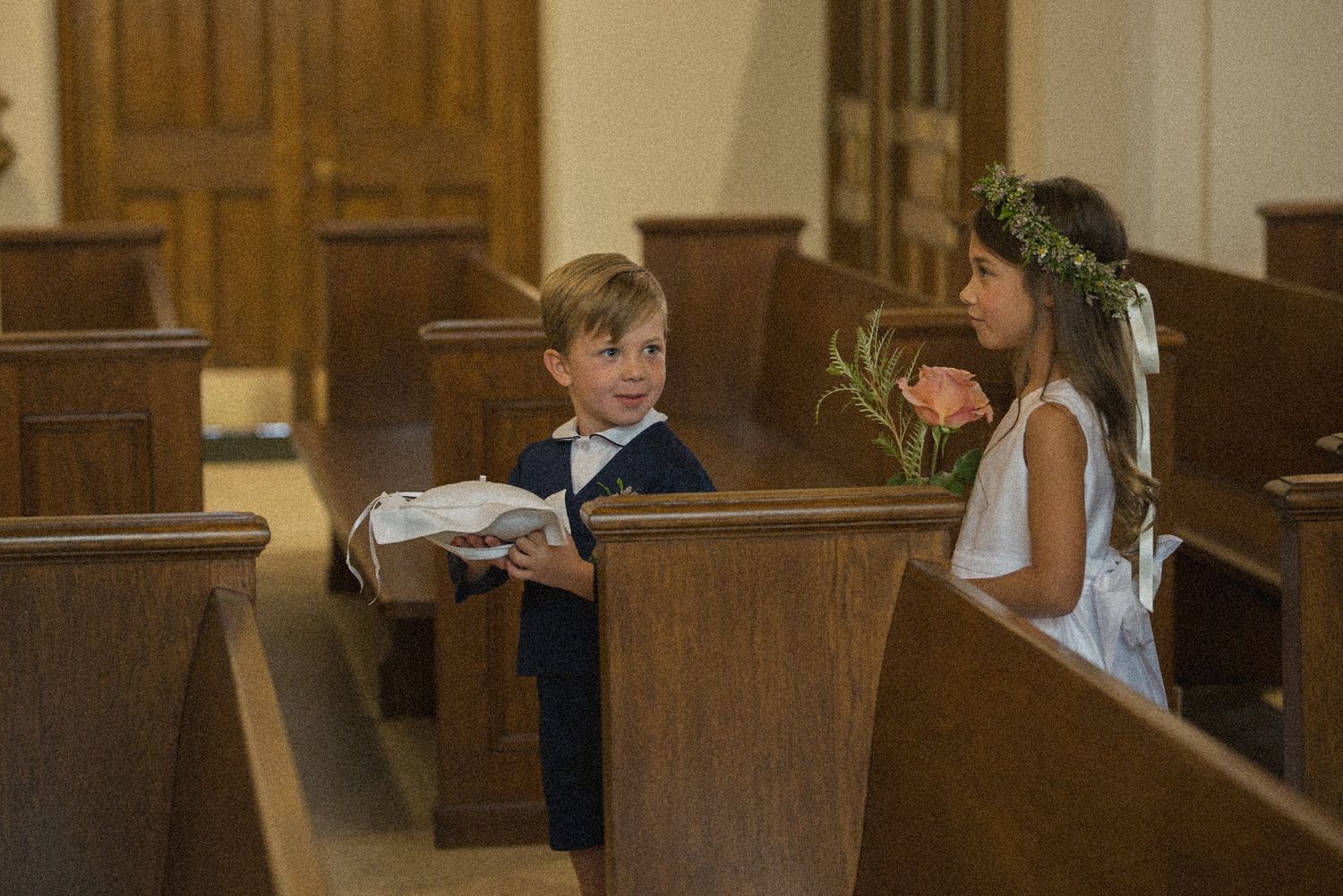 A young ring bearer in a navy suit holds a white pillow beside a flower girl wearing a floral crown between wooden church pews.