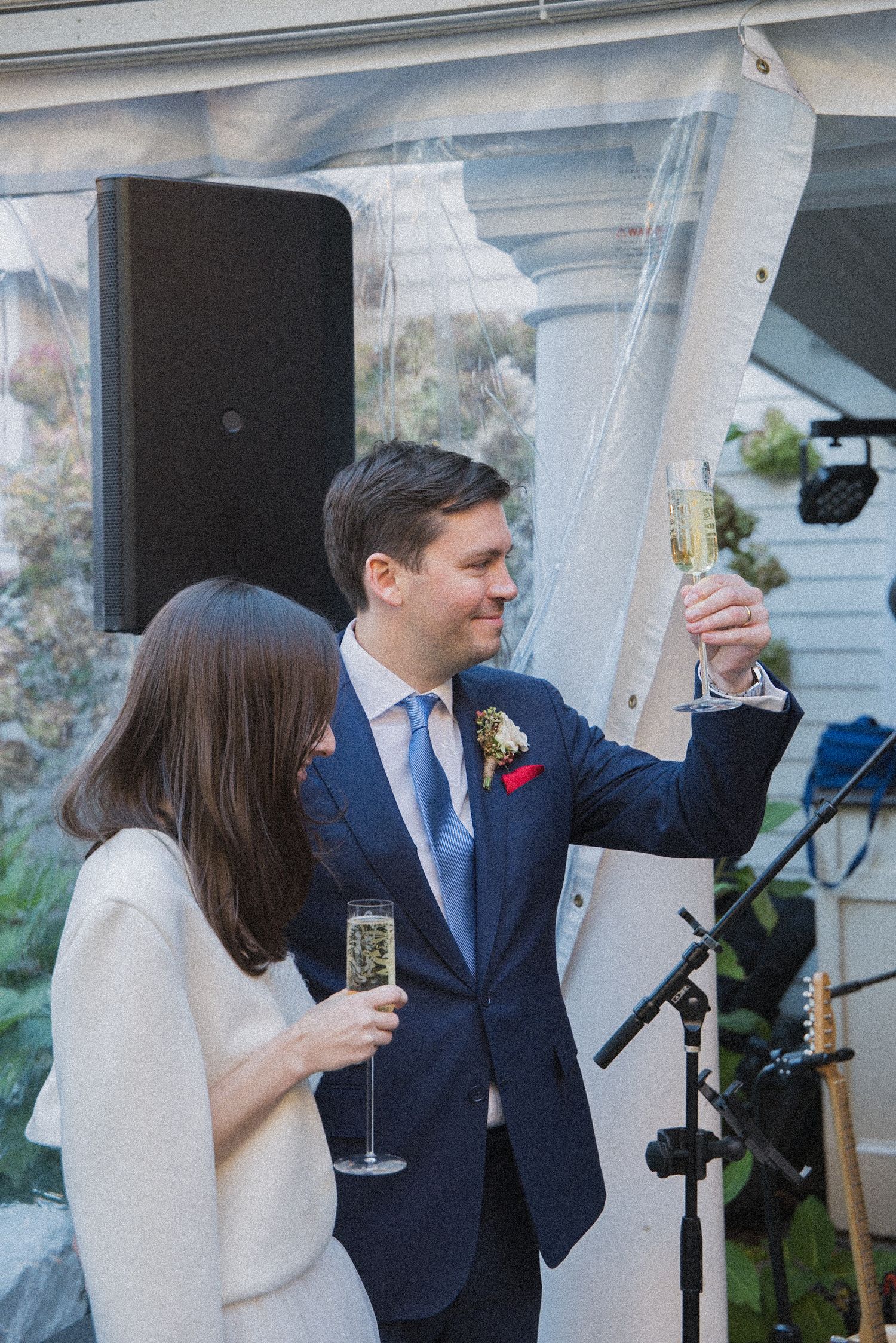 Groom in a navy suit with a floral boutonniere raises a champagne glass in a toast beside a woman holding a flute under a tent.