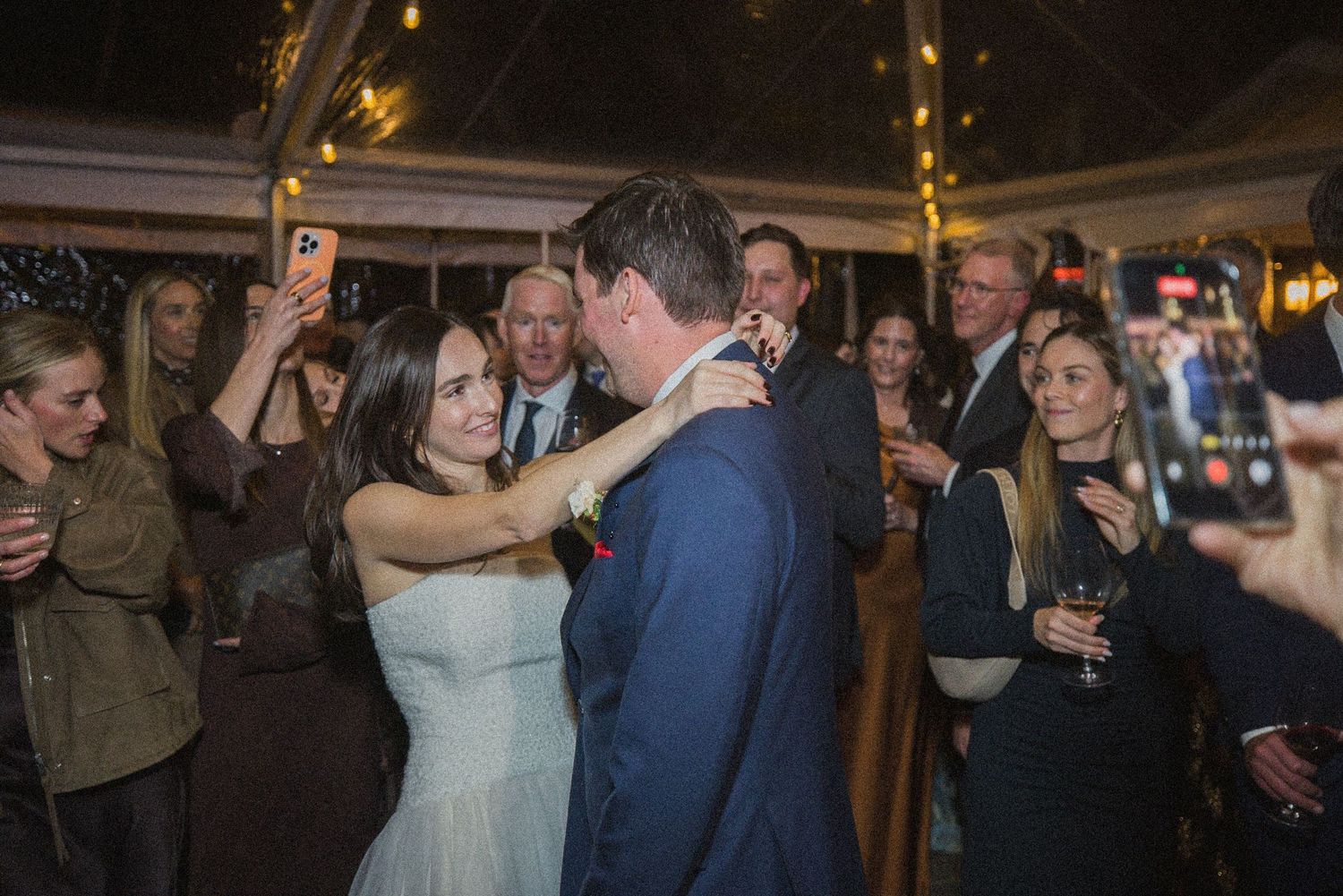 Bride in a white dress and groom in a navy suit share a dance under a tent as smiling wedding guests watch and take photos.