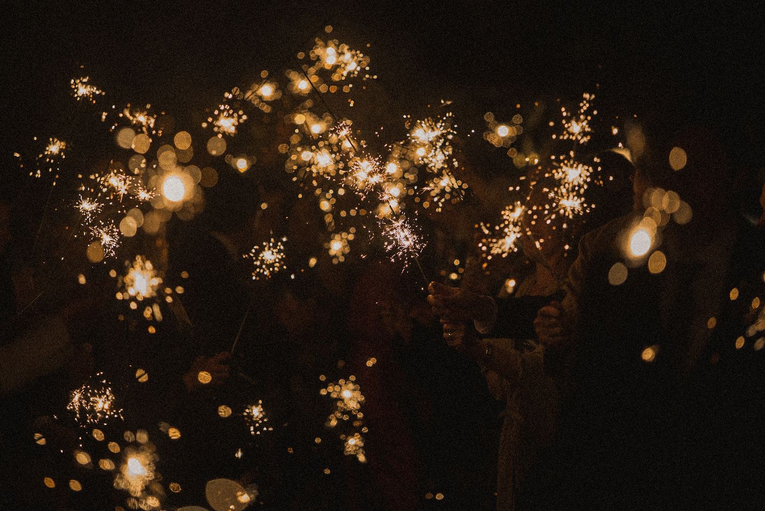 Glowing sparklers burning in the dark with golden sparks and soft bokeh lights, held by a crowd during a night celebration.