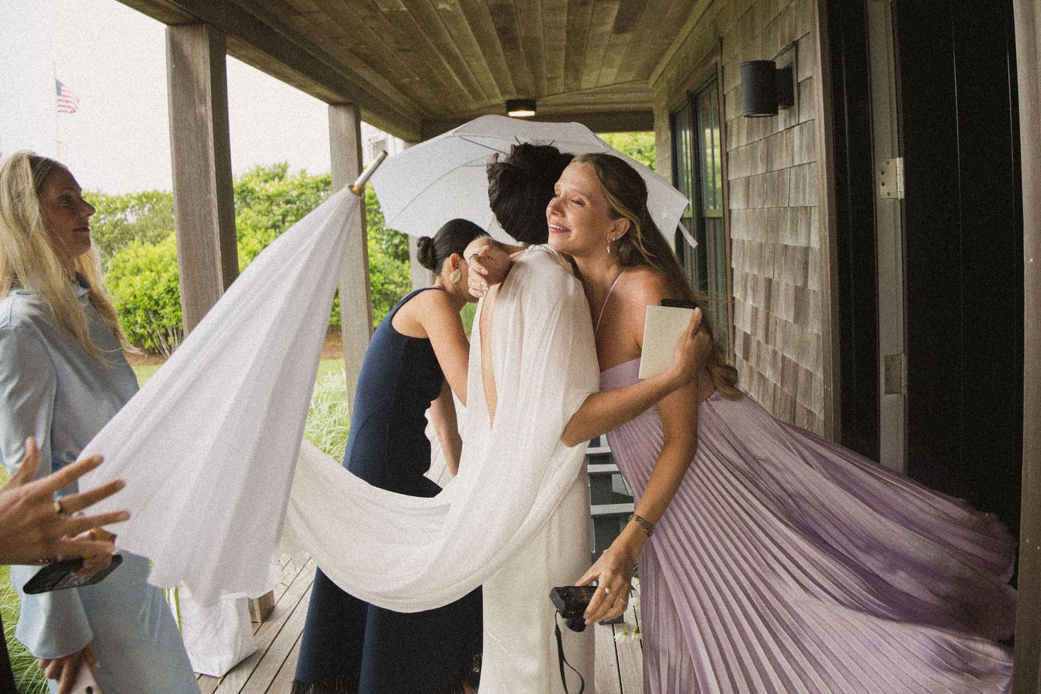 Bride in white gown and veil embraced by bridesmaids in lavender dresses on a wooden porch.