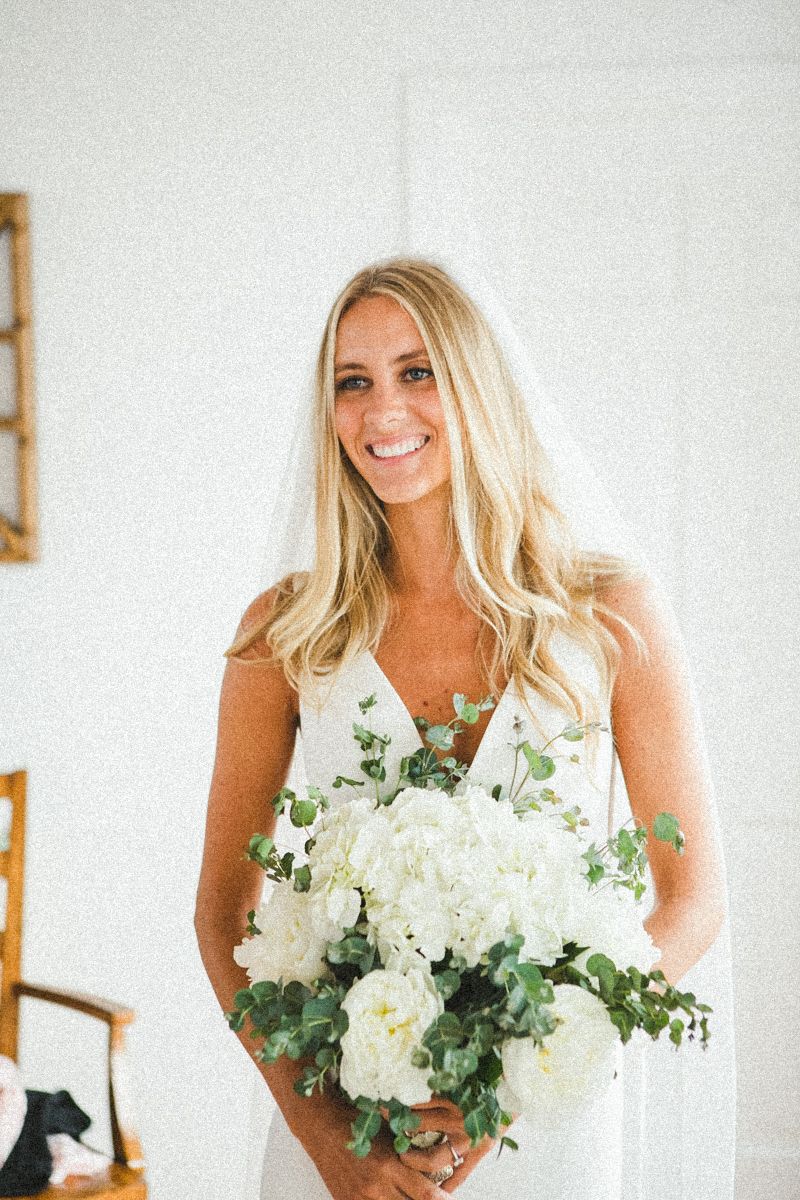 Person in white dress holding elegant white and green floral bouquet against white background.